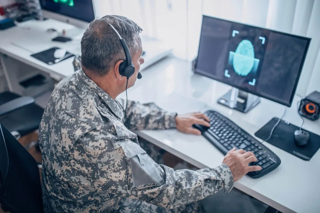 A senior military officer in camouflage uniform using a computer with two screens, displaying graphical data, in an office setting.